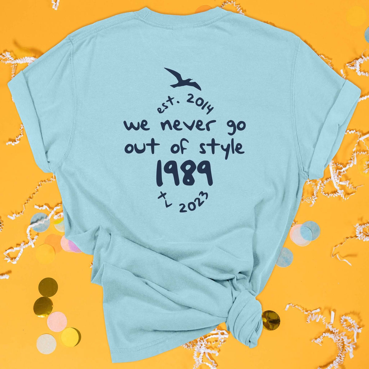 On a sunny mustard background sits the back of a t-shirt with white crinkle and big, colorful confetti scattered around. This Taylor Swift Inspired Reputation tee is aqua with navy lettering and an illustration of a seagull. It says "we never go out of style 1989" and "est. 2014 tv 2023" in handwritten lettering.