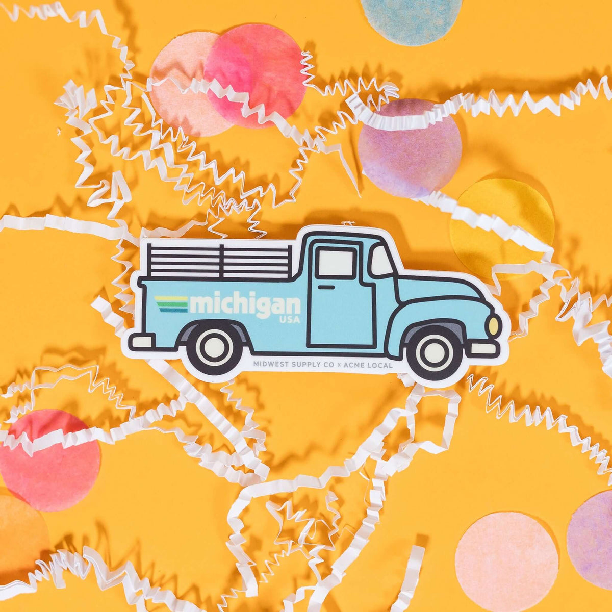 On a sunny mustard background sits a sticker with white crinkle and big, colorful confetti scattered around. This white sticker has an illustration of a light blue truck with black outlines and it says "michigan USA" in white block lettering with three stripes. The stripes colors are yellow, green, and blue. 2"-3"