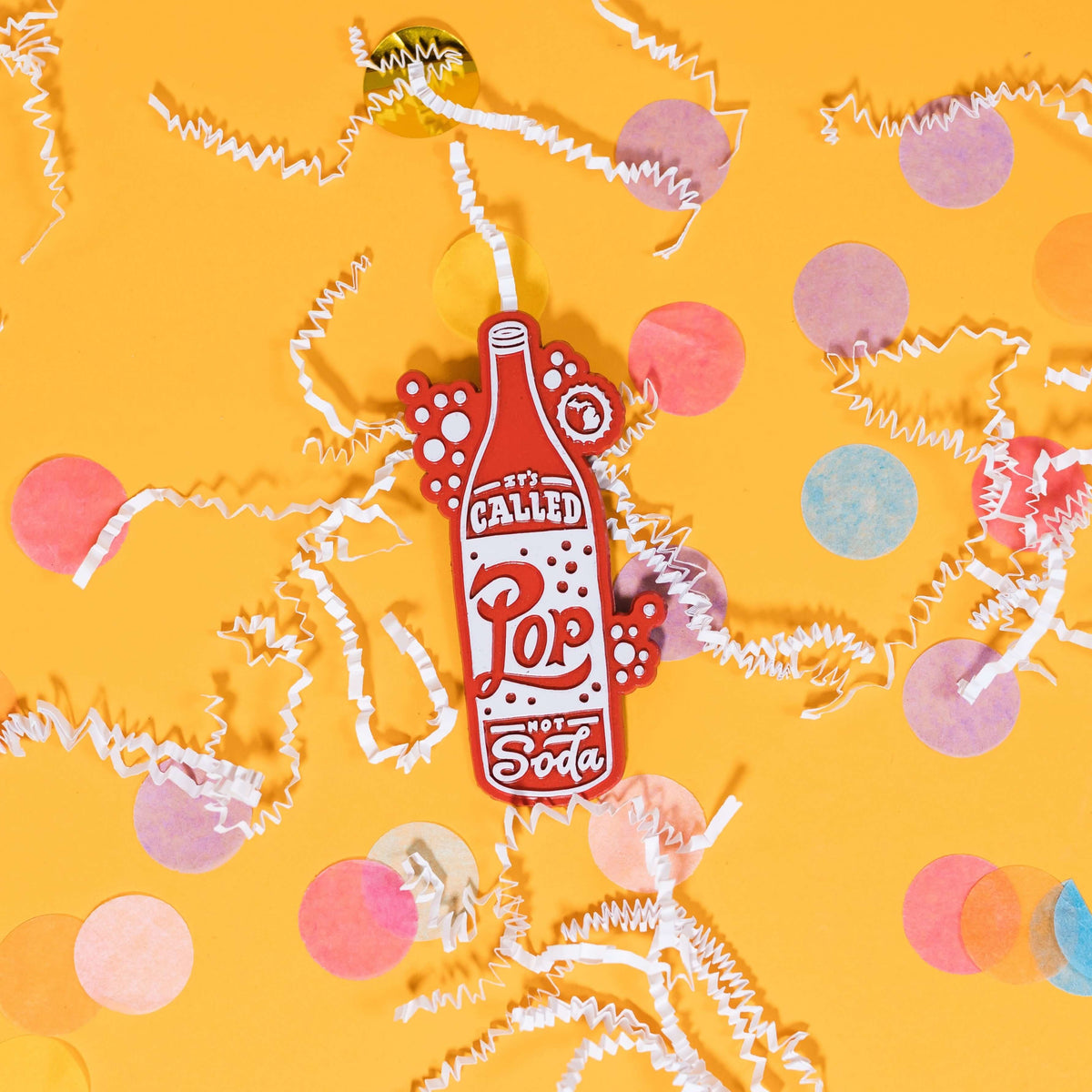 On a sunny mustard background sits a magnet with white crinkle and big, colorful confetti scattered around. This 2D-style raised magnet is a red and white handdrawn soda pop that says "It's Called Pop Not Soda" in red and white handwritten lettering. It has a white outline bubble with the state of Michigan in it in white. 3"- 4"