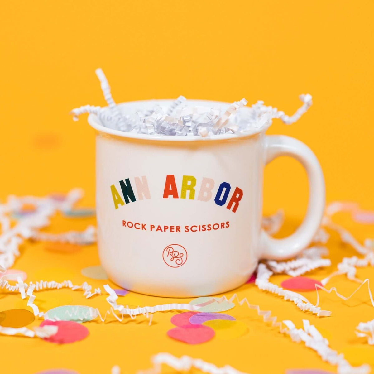 On a sunny mustard background sits a mug with white crinkle and big, colorful confetti scattered around. The white mug has a color collegiate font that says "ANN ARBOR" and under it says "ROCK PAPER SCISSORS" in a red, all caps block font. Under that is a circular, red "RPS" logo in handwritten script lettering.