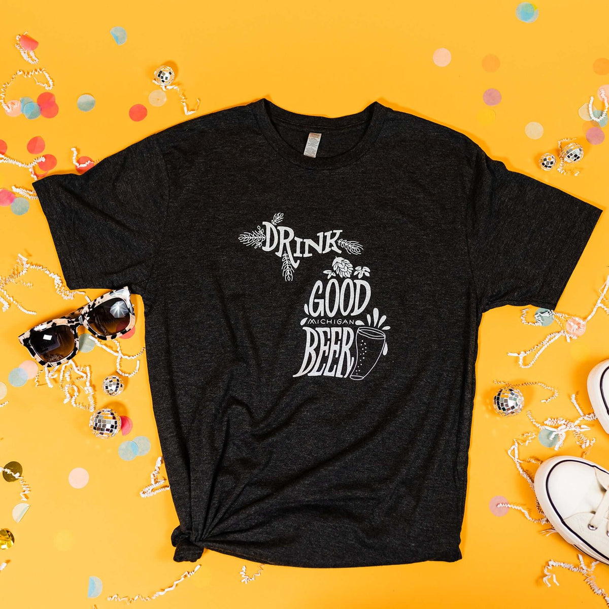 On a sunny mustard background sits a t-shirt with white crinkle and big, colorful confetti scattered around. There are mini disco balls, sunglasses, and a pair of white sneakers. This heathered charcoal grey tee features an illustration of a beer with hops and hand lettered "DRINK GOOD MICHIGAN BEER" in white.