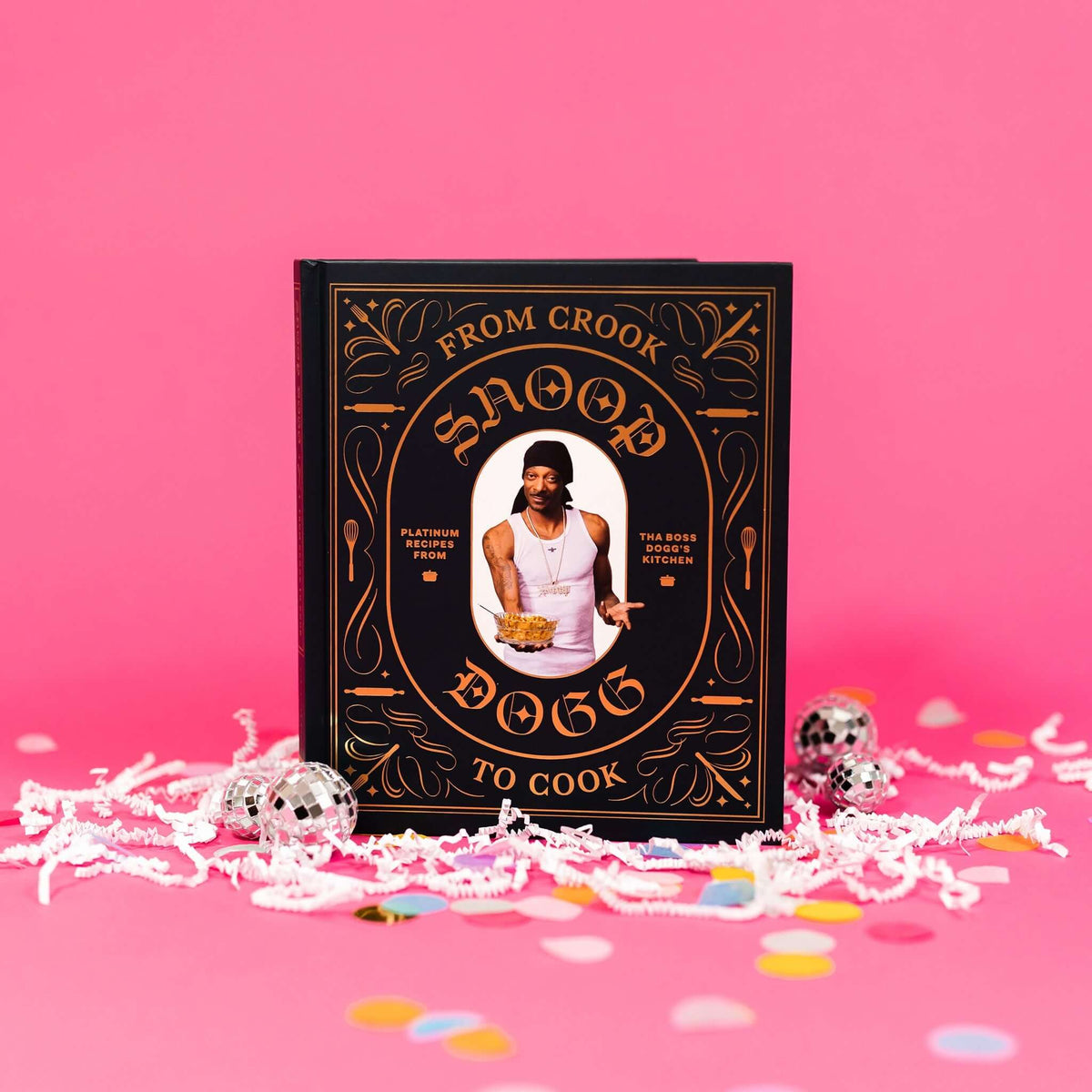 On a bubblegum pink background sits a book with white crinkle and big, colorful confetti scattered around. There are mini disco balls. This black cookbook has orange illustrations of kitchen utensils and a picture of Snoop Dogg holding a bowl of food. It says "FROM CROOK TO COOK SNOOP DOGG" and "PLATINUM RECIPES FROM THA BOSS DOGG'S KITCHEN."