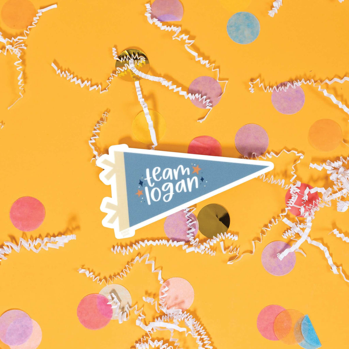 On a sunny mustard background is a sticker with white crinkle and big, colorful confetti scattered around. This Gilmore girls inspired vintage banner shaped sticker is new blue and khaki and has navy and orange stars. It says "team logan" in white, lower case handwritten lettering. 3"x1.6"