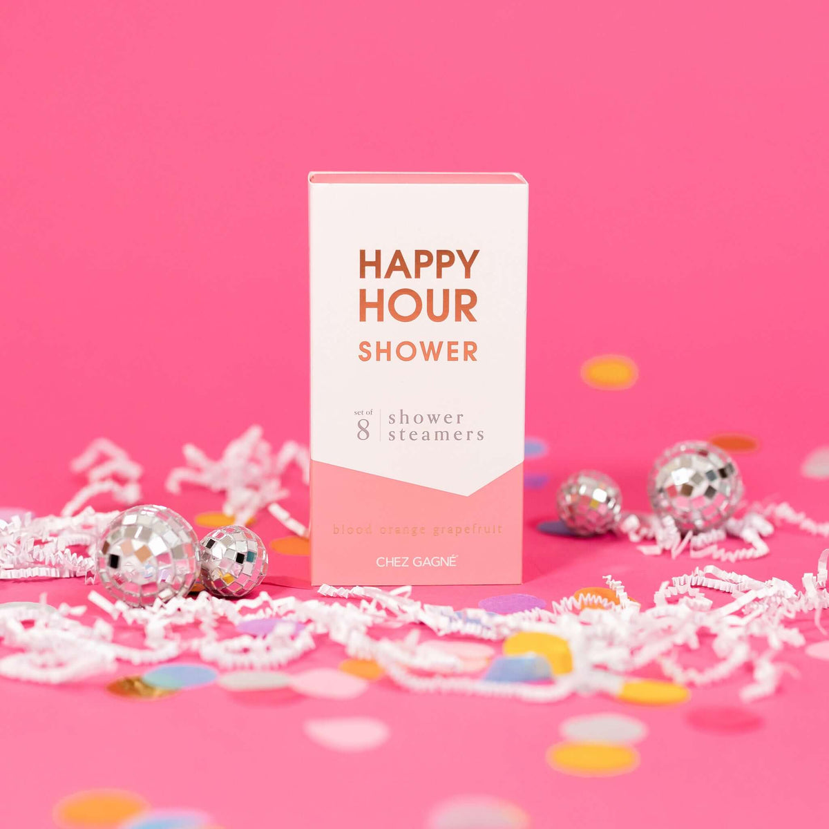 On a hot pink background sits a box with white crinkle and big, colorful confetti scattered around. There are mini disco balls. This picture is a close-up of a white and coral package that says "HAPPY HOUR SHOWER" in gold foil, all caps block lettering. Under it says "set of 8" and " shower steamers" in grey, lowercase serif font. At the bottom it says "blood orange grapefruit" in gold foil, lower case serif font. 