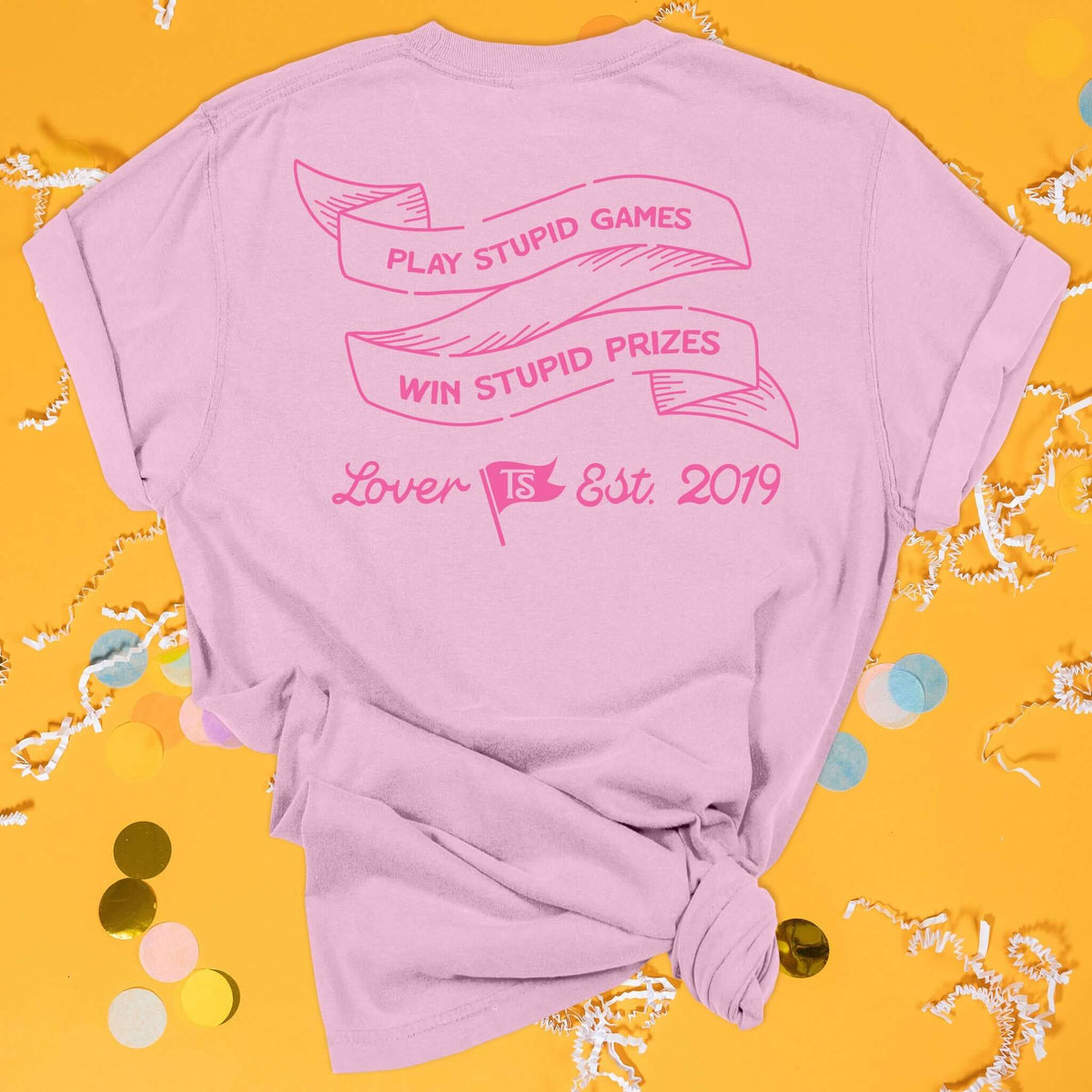 On a sunny mustard background sits the back of a t-shirt with white crinkle and big, colorful confetti scattered around. This Taylor Swift Inspired Reputation tee is pink with bubblegum lettering and ribbon illustration. In the ribbon it says "PLAY STUPID GAMES WIN STUPID PRIZES" in all caps. Under it says "Lover Est. 2019" in script lettering. There is a flag that has "TS" in it. 