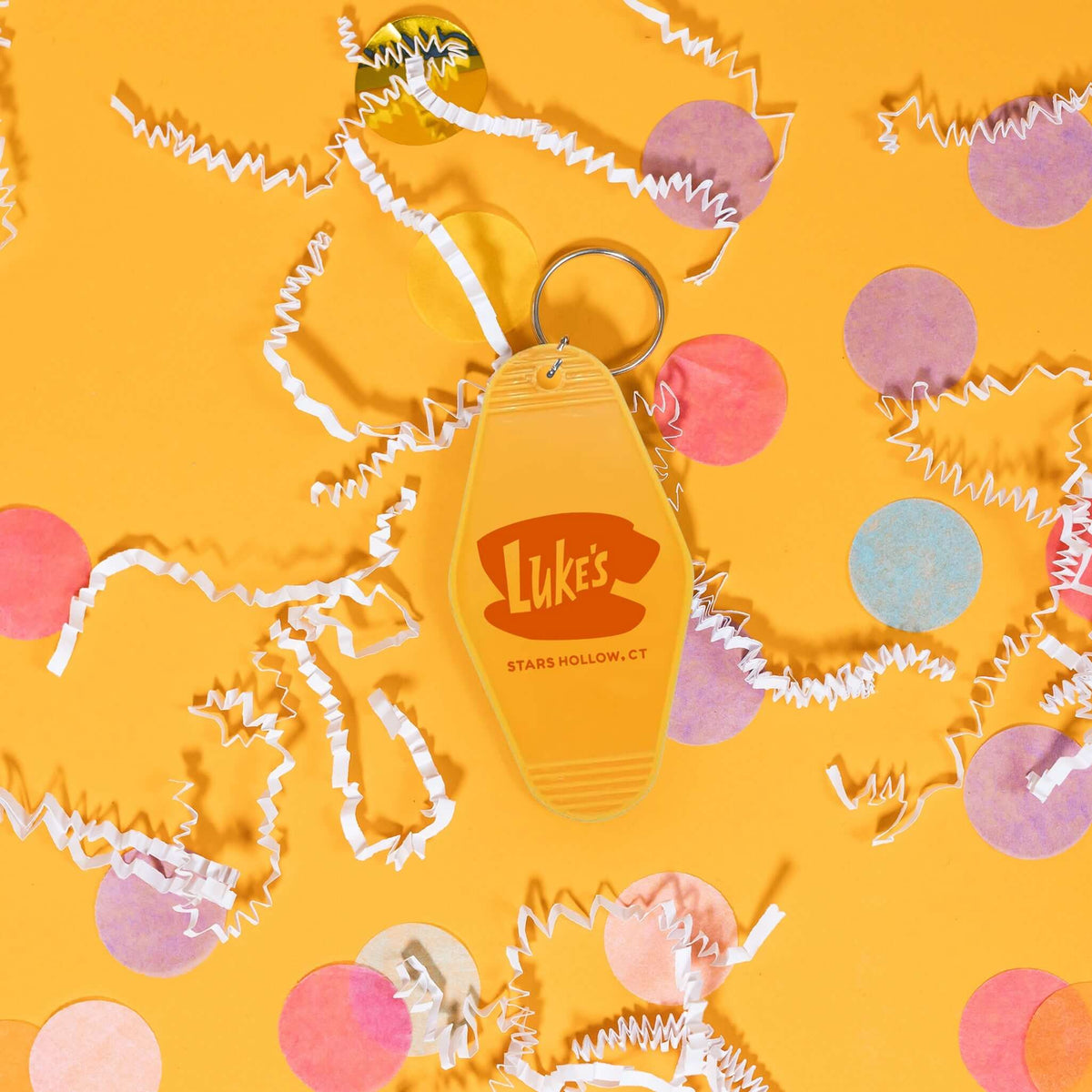 On a sunny mustard background sits a key chain with white crinkle and big, colorful confetti scattered around. This Gilmore Girls inspired mustard yellow vintage hotel key chain has a red illustration of Luke's Stars Hollow.