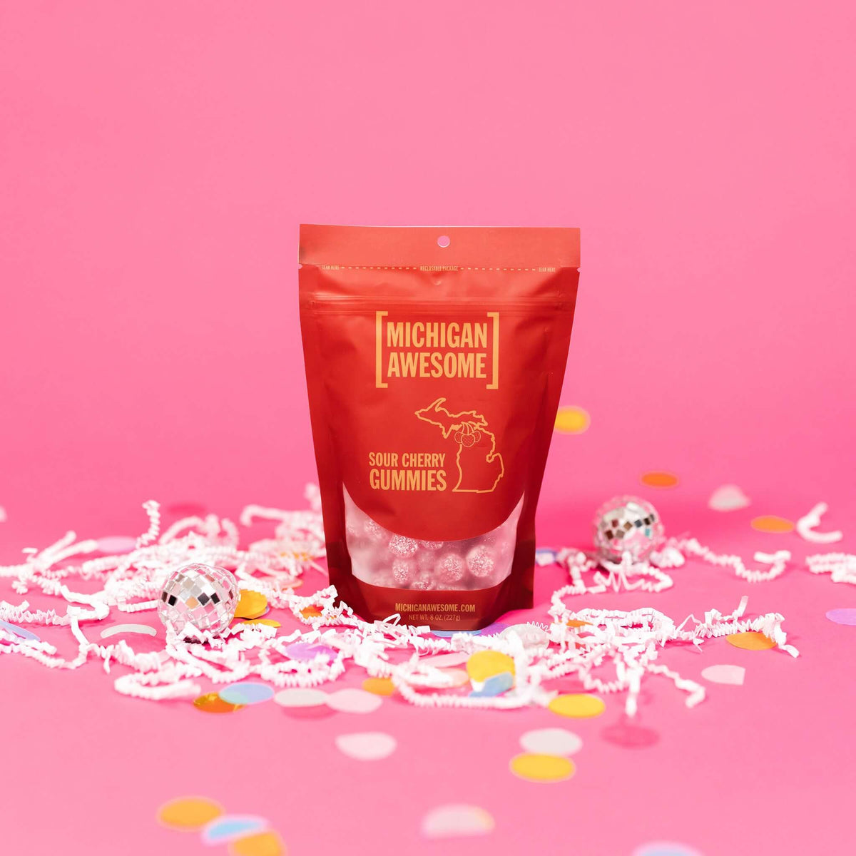 On a bubblegum pink background sits a bag with white crinkle and big, colorful confetti scattered around. There are mini disco balls. This red bag filled with sugar sour cherry gummies. It says "MICHIGAN AWESOME," and "SOUR CHERRY GUMMIES" in goldish-yellow, all caps block font. It has an outlined illustration of the state of Michigan-UP with cherries. There are sugar sour cherry gummies sitting around the bag.