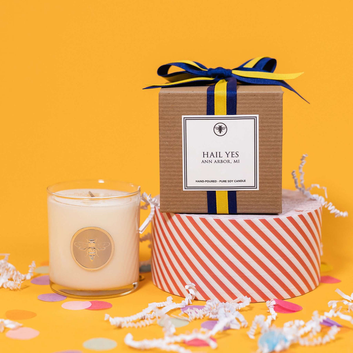 On a sunny mustard background is a candle with a box and white crinkle with big, colorful confetti scattered around. The clear glass candle has a gold foil round label with an illustration of a bee on it. There is a kraft box with yellow and navy striped ribbon tied at the top and on the front is a square white label that says "Hail Yes Ann Arbor Mi" and it sits atop a red and white striped packing tape. The candle is a "Hand-poured, pure soy candle."