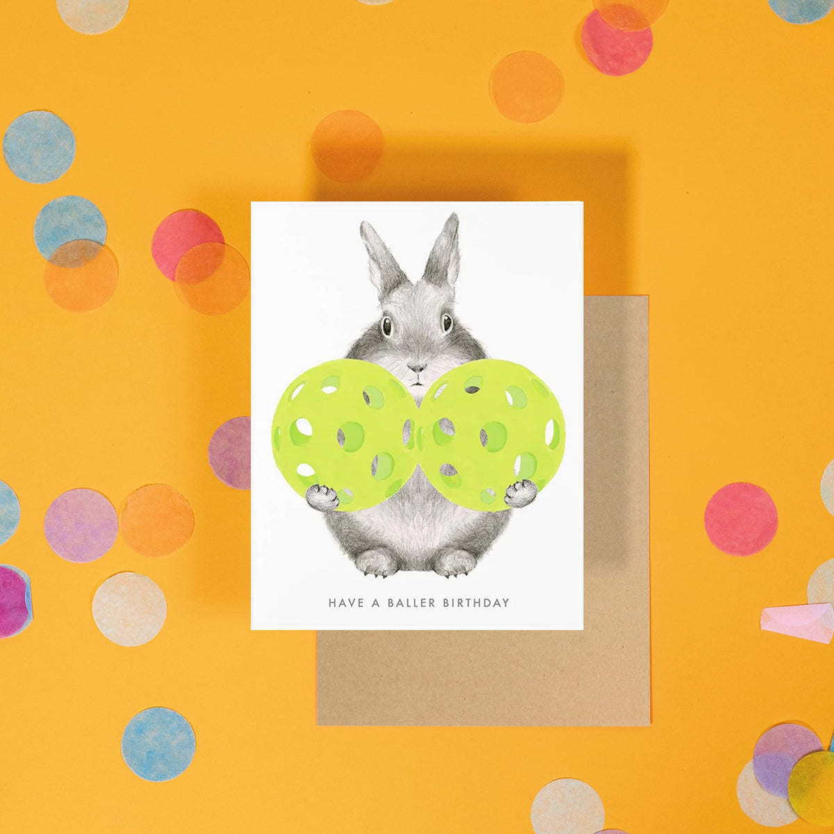 A greeting card sits on a bright yellow background with colorful confetti with a hand drawn bunny with hand painted pickleballs text reads "Have a Baller Birthday". 