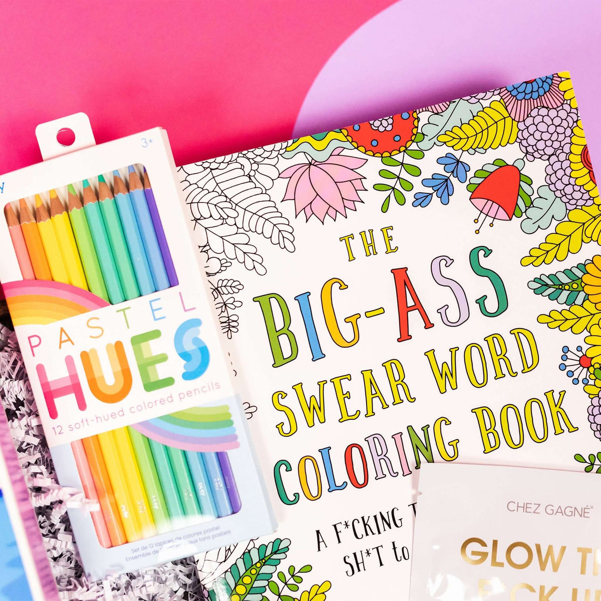 On a bright pink and purple background, a handful of optimistic, brightly colored swear-themed gifts are arranged on a tray among white crinkle. The photo is a close-up of the 'Big-Ass Swear Word Coloring Book' with flowers and Ooly Pastel hues colored pencils.