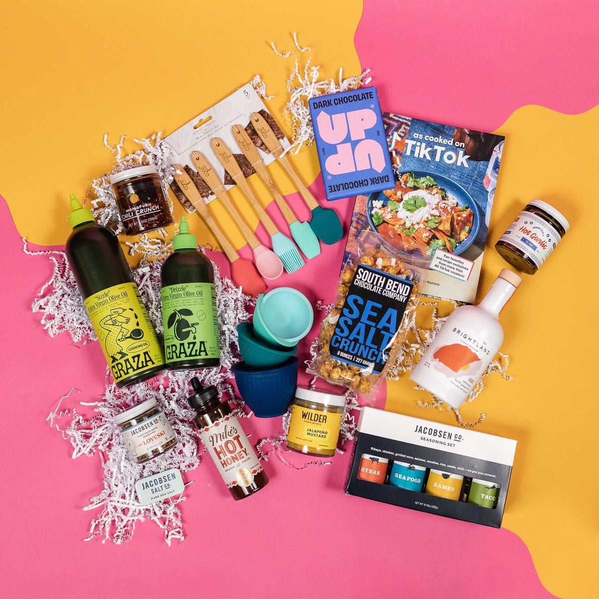 On a hot pink background with mustard wobbly cutout shapes, an assortment of lux kitchen accoutrements are arranged on white paper crinkle. The gifts include two Graza olive oils, multicolored silicone utensils, Jacobson Salt Co. flavored salts, Mike's Hot Honey, "As Cooked on TikTok" cookbook, Momofuku Chili Crunch, Silicone finger bowls, Wilder Jalapeño Mustard, Brightland Olive Oil, Mama Teay's Hot Garlic, Up Up Dark Chocolate and South Bend Popcorn Company Sea Salt Crunch Caramel Corn.