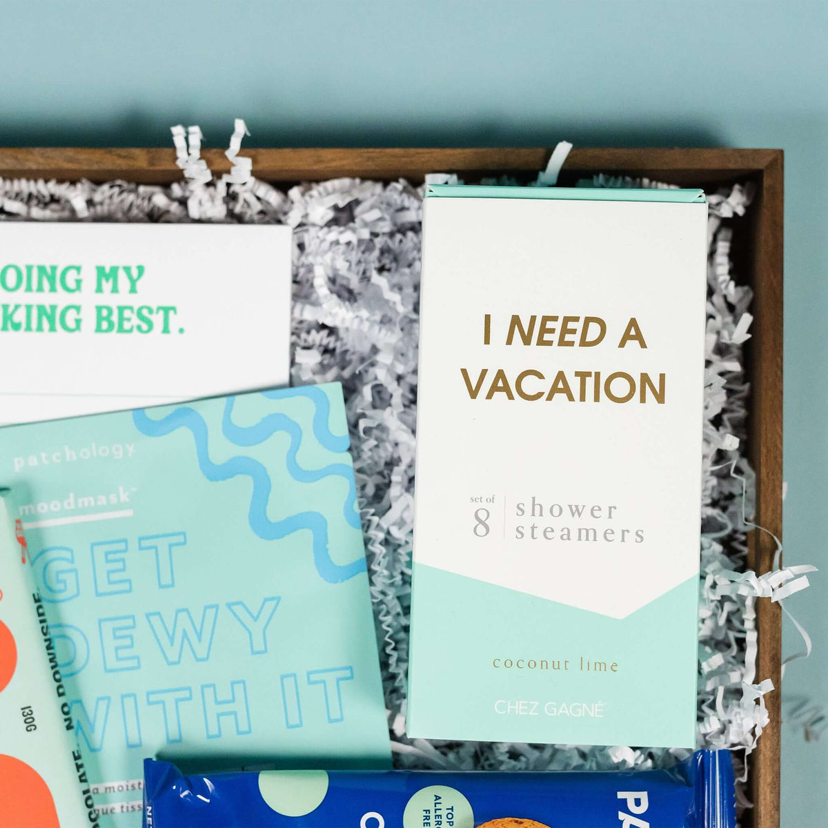 On a calming blue background sits a wooden tray of products and white paper krinkle. The products are a lovely sky blue and aqua theme. This photo is a close-up on the 8 pack of Chez Gagne coconut lime shower steamers that say "I Need a Vacation" and  Patchology moisturizing sheet mask "Get Dewy With It."