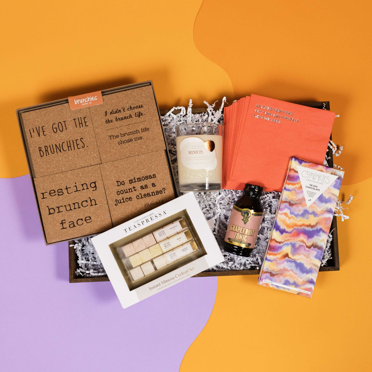 On a funky light orange and lavender background sits a wooden tray filled to the brim with brunch accoutrements. The products include a set of cork brunch-themed coasters, our favorite Teaspressa Instant Mimosa Cocktail Kit, a premium 100% soy Mimosa candle, a small bottle of Portland Syrups Grapefruit Tonic drink mixer, a stack of coral beverage napkins that say "STARTED FROM THE BOTTOMLESS MIMOSAS NOW WE HERE," and a gorgeous Compartes Tie Dye White Chocolate Bar.