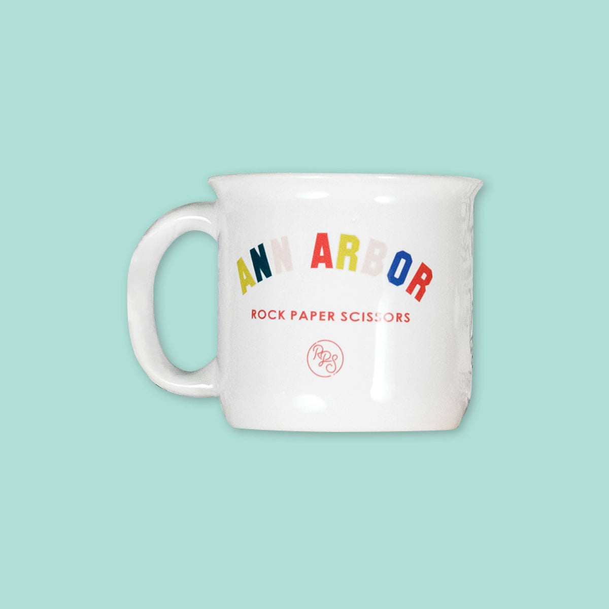 On a mint green background sits a mug. The white mug has a color collegiate font that says "ANN ARBOR" and under it says "ROCK PAPER SCISSORS" in a red, all caps block font. Under that is a circular, red "RPS" logo in handwritten script lettering.
