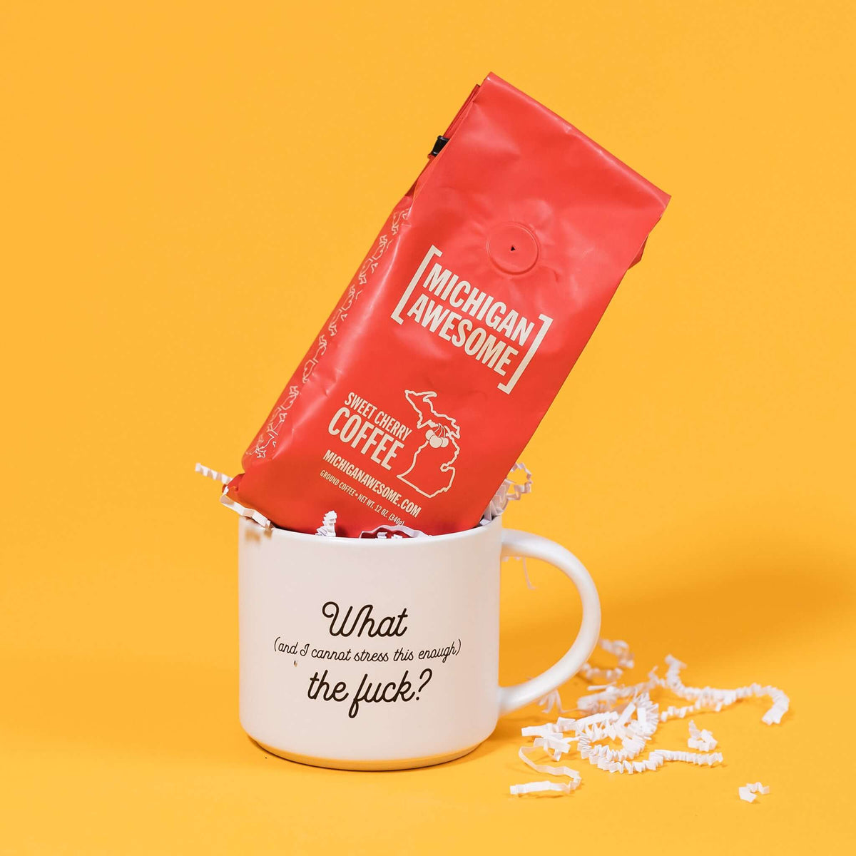 On a mustard yellow background is a photo of a mug and a bag of coffee. The mug is a white diner style mug that says in black script, "What (and I cannot stress this enough) the fuck?" A red bag of Michigan Awesome Sweet Cherry ground coffee sits atop the mug. White crinkle also overflows from the mug.