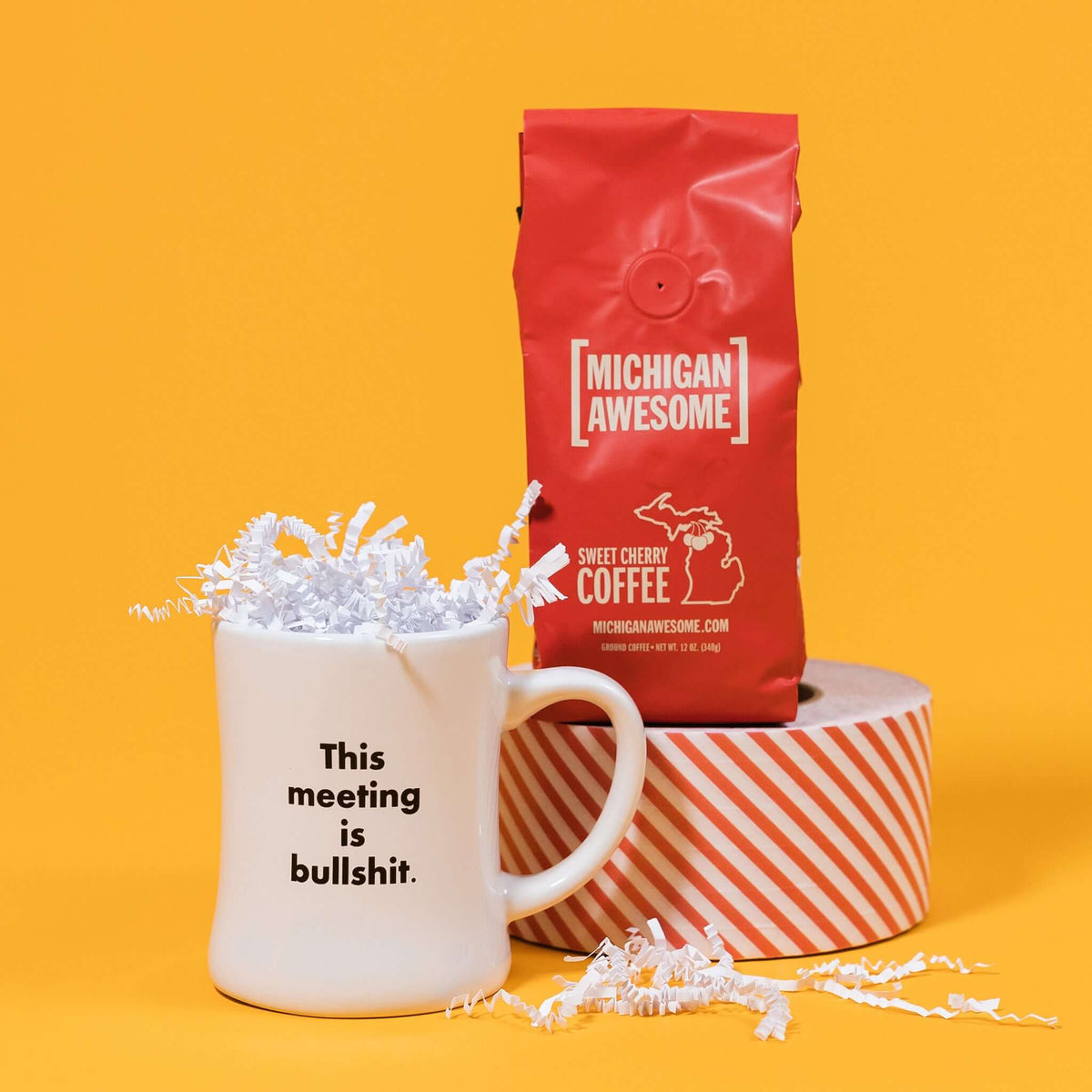 On a mustard yellow background is a photo of a mug and a bag of coffee. The mug is a white diner style mug that says in black basic type, "This meeting is bullshit." A red bag of Michigan Awesome Sweet Cherry ground coffee sits atop a roll of red and white striped tape. White crinkle overflows from the mug.