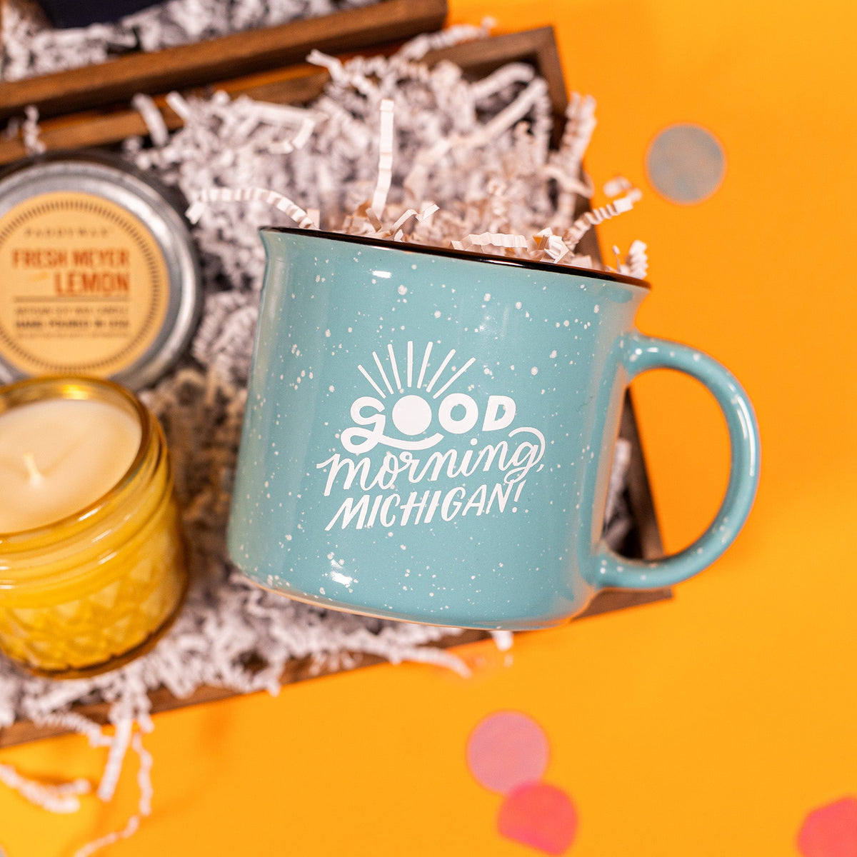 On a bright yellow background with coral pink confetti sits a gift box with white crinkle paper and a 15 oz. light teal campfire mug that says "Good Morning Michigan" in white writing and a 4 oz. yellow glass Paddywax Fresh Meyer Lemon candle with the lid removed.