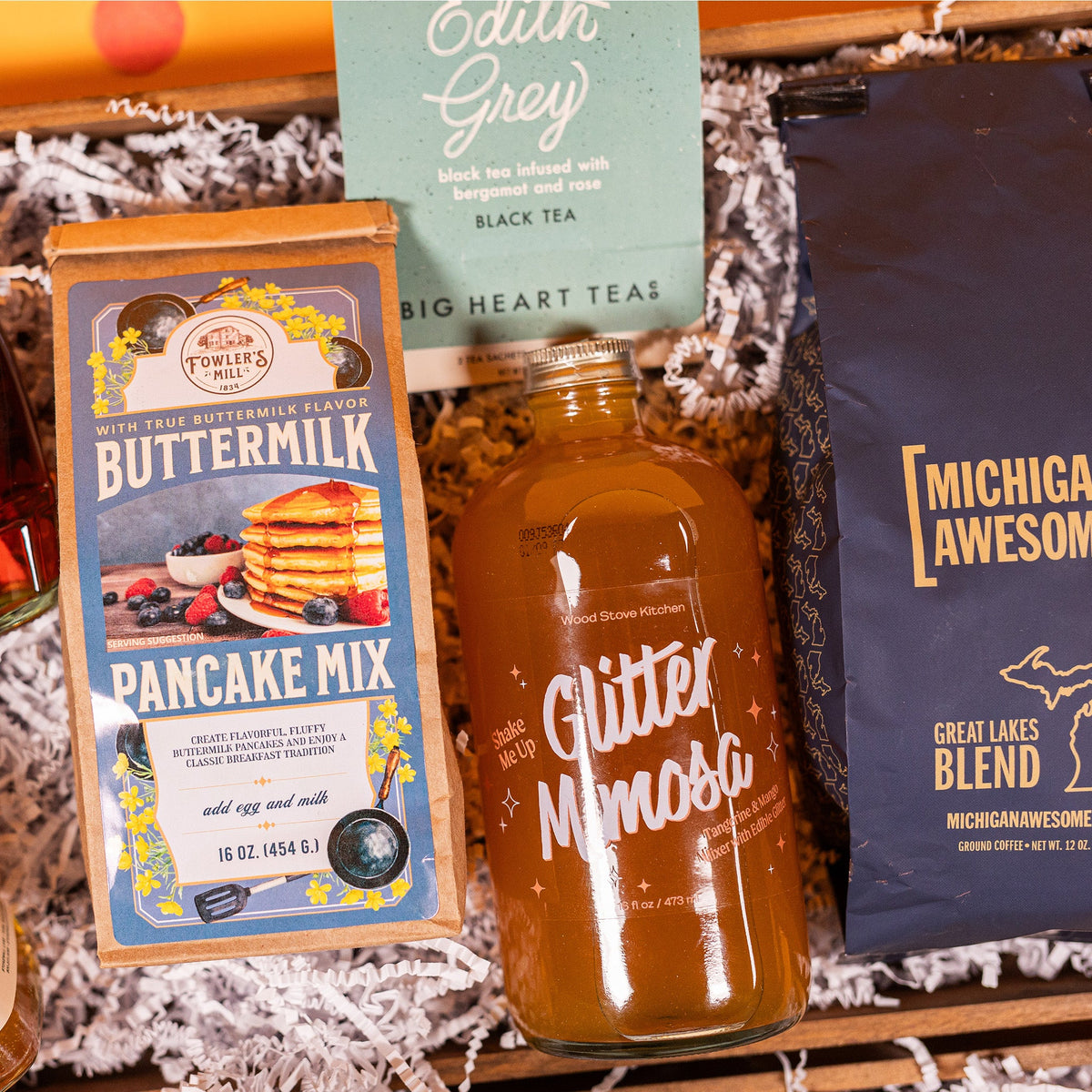 On a bright yellow background with coral pink confetti sits a gift box with white crinkle paper and a 16 oz.  bag of Fowler's Mill Pancake Mix, a teal and white box of Edith Grey tea from Big Heart Tea with two satchets, a 16 oz. glass bottle of Tangerine and Mango Glitter Mimosa beverage from Wood Stove Kitchen, and a 12 oz. bag of Great Lakes Blend coffee from Michigan Awesome. 