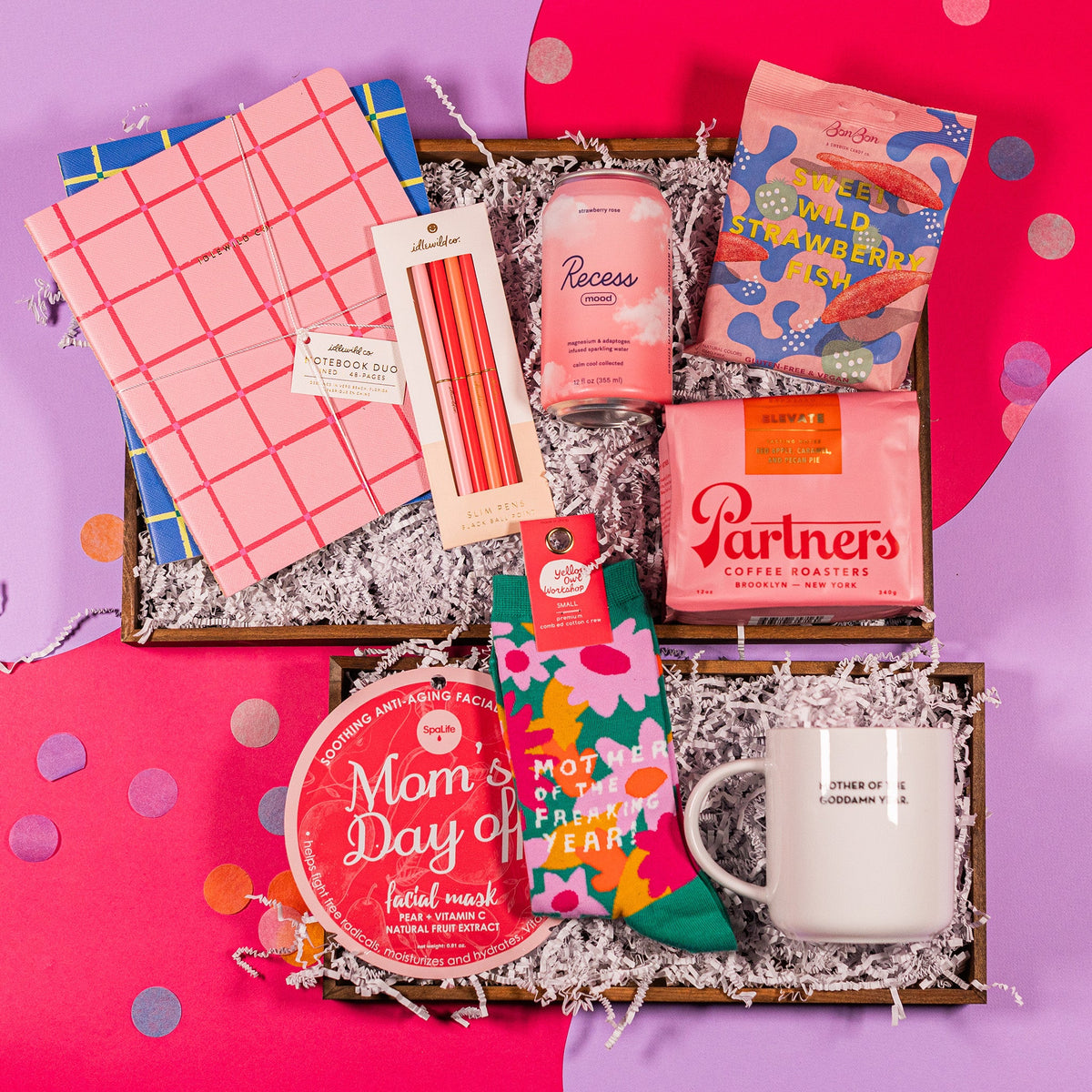 On a bright pink and lavender background with colorful confetti sits a collection of items in a gift box with white crinkle paper. A set of Slim Pink Pens, two notebooks, a can of Strawberry Rose beverage, a pouch of Sweet Wild Strawberry Fish candies, a pouch of Elevate Whole Bean Coffee, a pair of dark teal socks that say "Mother of the Freaking Year!", a white coffee mug that says "Mother of the Goddamn Year" in small black type, and a package containing a Mom's Day Off Facial Mask. 