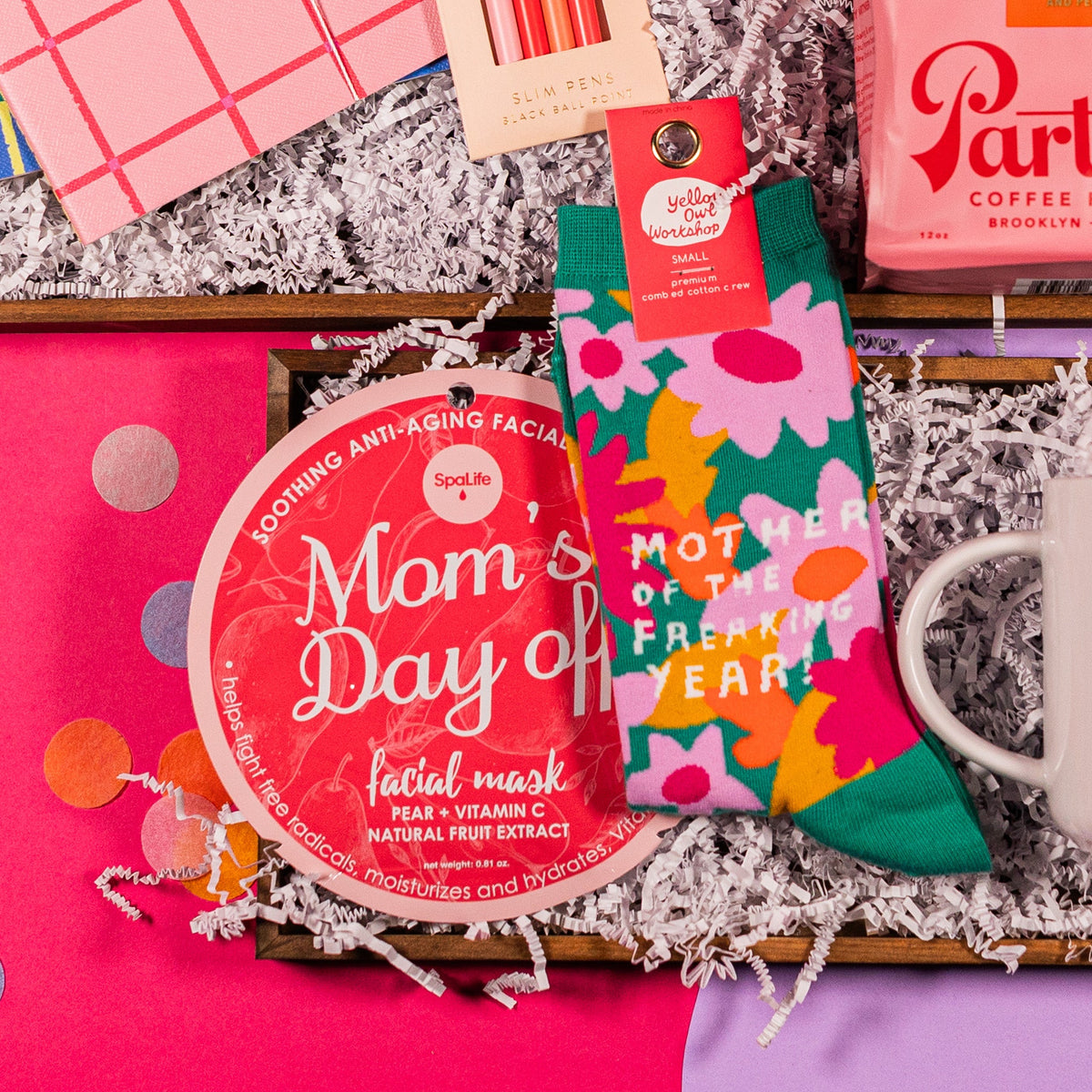 On a bright pink and lavender background with colorful confetti sits gift box and white crinkle paper with a pair of dark teal socks with pink and yellow flowers that say "Mother of the Freaking Year!" in white writing and a dark pink circular package containing a Mom's Day Off Facial Mask from Spa Life in gift box with white crinkle paper.