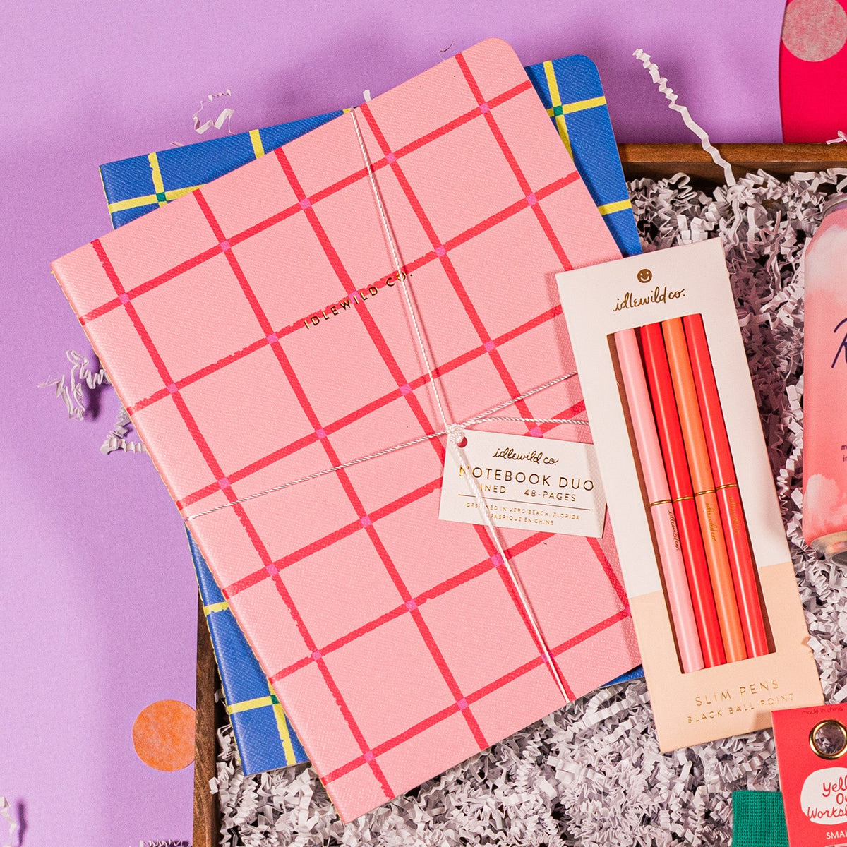 On a lavender background with colorful confetti sits a gift box and white crinkle paper with a 4-count set of Idlewild Co. Slim Pink Pens and two notebooks with 48 pages each, the top one pink with darker pink plaid stripes and the bottom one blue with yellow plaid stripes from Idlewild Co.