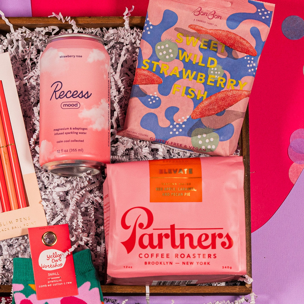 On a bright pink and lavender background with colorful confetti sits a gift box and white crinkle paper with a pouch of Sweet Wild Strawberry Fish candies from BonBon, a pink 12 oz. pouch of Elevate Whole Bean Coffee from Partners Coffee Roasters and a 12 fl. oz. pink can of Strawberry Rose beverage from Recess.