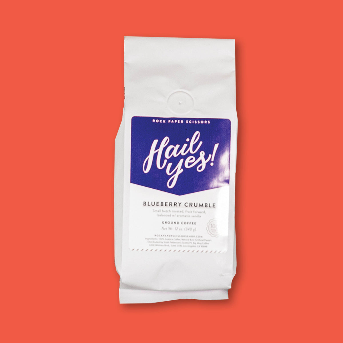 On a tomato red background sits a a white bag with blue label that says "Hail Yes!" containing Blueberry Crumble coffee from Rock Paper Scissors.