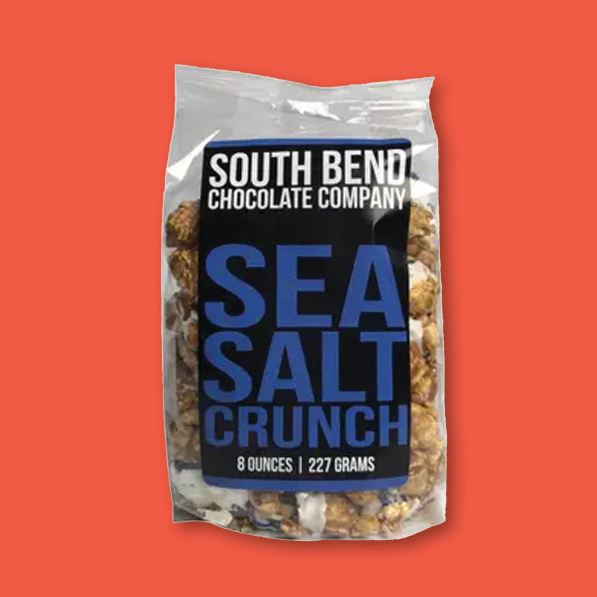 On a tomato red background sits a clear 8 oz. bag of Sea Salt Crunch Popcorn from South Bend Chocolate Company.