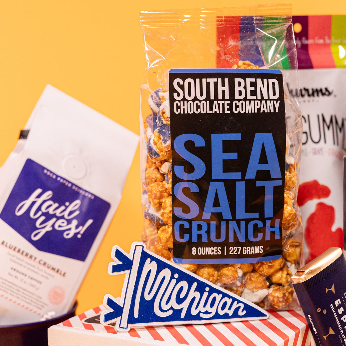 On a bright yellow background sits a clear 8 oz. bag of Sea Salt Crunch Popcorn from South Bend Chocolate Company, an 8 oz. white and rainbow colored pouch of Michi-gummies from Shurms Candy, a white bag with blue label that says "Hail Yes!" of Blueberry Crumble coffee from Rock Paper Scissors, a small blue pennant shape sticker that says Michigan in white script writing. 