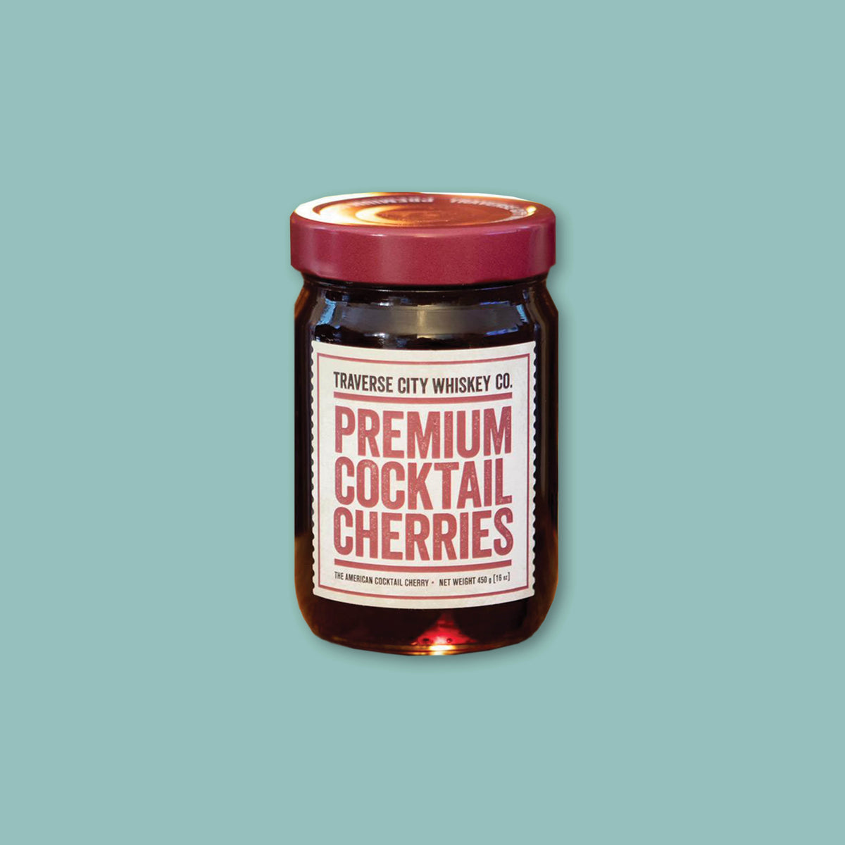 On a teal background sits a jar of Premium Cocktail Cherries from Traverse City Whiskey Co. with a white label.