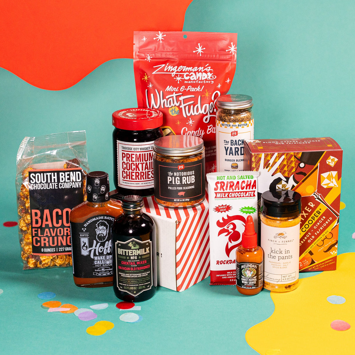 On an orange, teal, and yellow background with colorful confetti sits a collection of items. A box of Infused Ice Cubes, a bag of Bacon Flavored Crunch popcorn, a bag of What the Fudge Candy Bars, a Sriracha flavored chocolate bar, a jar of Kick in the Pants seasoning, a bottle of Hoff's Wakeup Call hot sauce, a bottle of Bittermilk Cocktail Mixer, a jar of Premium Cocktail Cherries, a jar of Burger Blend seasoning, a bottle of Melinda's Wing Sauce, and a jar of pork seasoning.