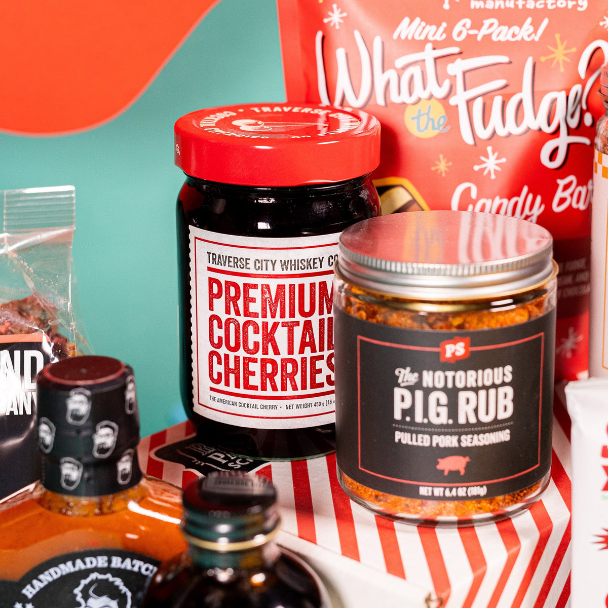 On a red and teal background sits a red bag of Zingerman's candy bars, a jar of The Notorious P.I.G. Run, and a jar of Premium Cocktail Cherries with a red lid.