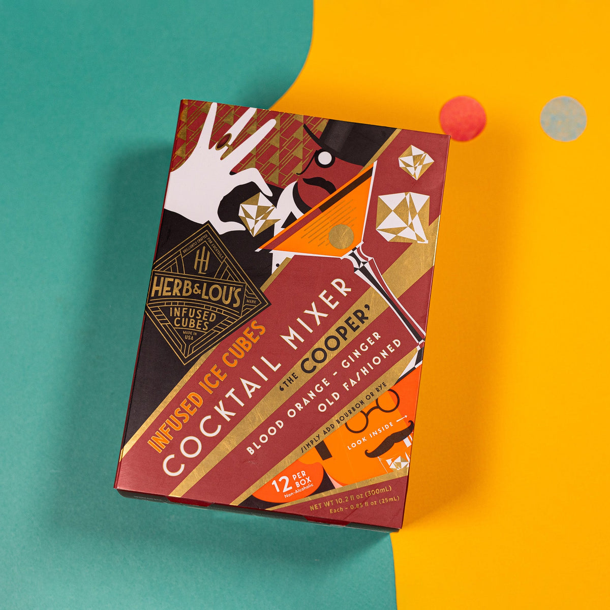 On a teal and yellow background sits a burgundy 12-count box of Herb & Lou's Infused Ice Cubes, Blood Orange and Ginger flavored.