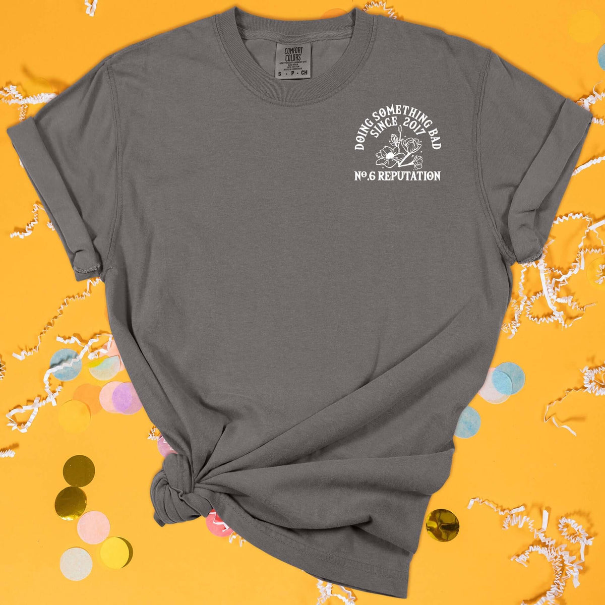 On a sunny mustard background sits the front of a t-shirt with white crinkle and big, colorful confetti scattered around. This Taylor Swift Inspired Reputation tee is dark grey with white lettering and illustration over the top left chest. There are flowers and it says "DOING SOMETHING BAD SINCE 2017, NO.6 REPUTATION" in all caps.