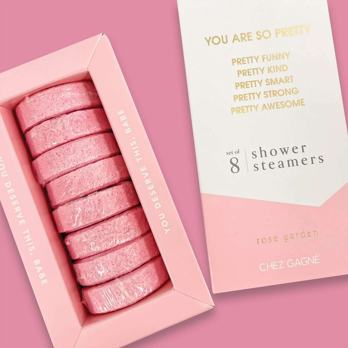 On a bubblegum pink background sits two boxes. This picture is a close-up of a white and light pink package that says "YOU ARE SO PRETTY" and "PRETTY FUNNY PRETTY KIND PRETTY SMART PRETTY STRONG PRETTY AWESOME" in gold foil, all caps block lettering. Under it says "set of 8" and " shower steamers" in grey, lowercase serif font. At the bottom it says "rosé garden" in gold foil, lower case serif font. To the left of it is a light pink box with pink shower steamers in it. 